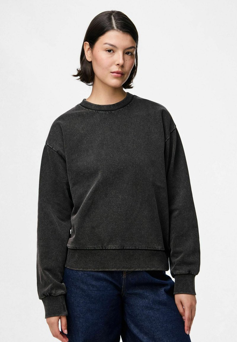 Jauna moteris su trumpais tamsiais plaukais, vilkinti nusipraususiu juodu ilgomis rankovėmis megztiniu ir tamsiai mėlynomis džinsomis, stovinti prieš paprastą šviesų foną.