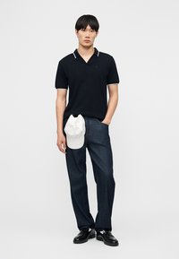 Νεαρός άνδρας στέκεται φορώντας μαύρο πόλο μπλουζάκι με λευκή διακόσμηση, σκούρο τζιν, μαύρα παπούτσια, κρατώντας ένα λευκό καπέλο στο ένα χέρι, μπροστά σε απλό φόντο.