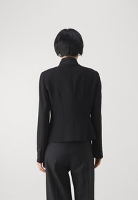 Blazer nero su misura con un design aderente, colletto a punta e spacco posteriore. Tessuto opaco con polsini con bottone e una silhouette strutturata.