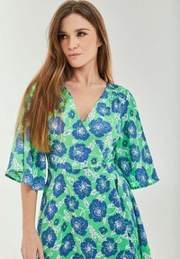 Floral wrap dress featuring a v-neck, green base with blue flowers, and short, flared sleeves. Made of lightweight fabric with a smooth texture.