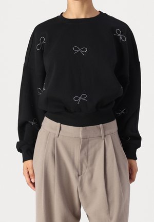 Zwart shirt met lange mouwen en kleine witte stippen in de vorm van strikjes, gecombineerd met hoog getailleerde beige geplooide broek, gedragen door een staand persoon.