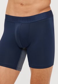 Navy blue boxer shorts featuring a textured gray side panel and a logo waistband. Made from a smooth, stretchy material for comfort.