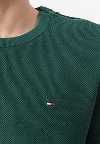 Pull en tricot vert avec un col rond et un détail zippé à l'épaule. Présente un petit logo rouge, blanc et bleu sur la poitrine.