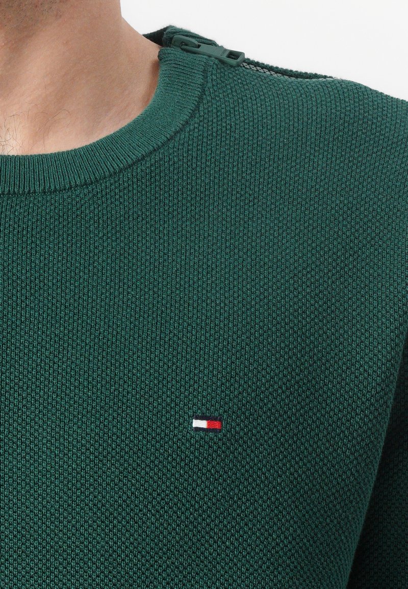 Groene gemêleerde trui met een ronde hals en een ritsdetail op de schouder. Heeft een klein rood, wit en blauw logo op de borst.