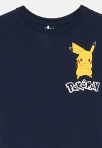 Tamsiai mėlynas medvilninis marškinėliai su geltonu Pikachu grafiniu vaizdu ir žodžiais "Pokémon" balta spalva po personažu. Trumpomis rankovėmis, apvaliu kaklu.