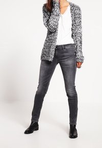 Svartvit stickad kofta över en vit t-shirt, kombinerad med figurnära grå jeans och svarta stövletter. Koftan har en texturerad design.