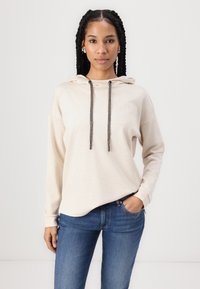 Sweatshirt bege com capuz, de corte solto e mangas longas. Apresenta um capuz com cordão e um tecido texturizado, combinado com jeans azuis.