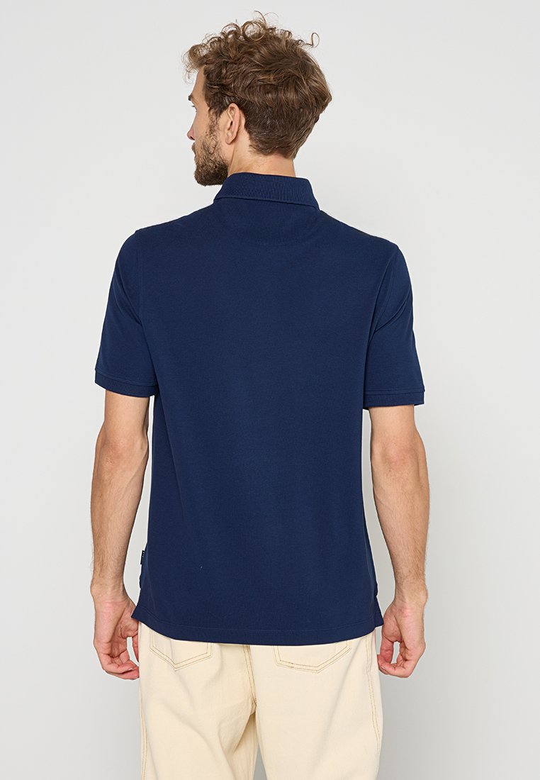 Homme avec des cheveux bruns bouclés portant un polo bleu marine à manches courtes et un pantalon beige, de dos contre un fond uni.