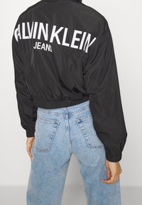 Černá zkrácená bunda na zip s velkým bílým nápisem "CALVIN KLEIN JEANS" na zádech; v kombinaci s světle modrými džíny s vysokým pasem.
