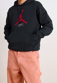 Persona vistiendo una sudadera negra con un logotipo de un jumper de baloncesto rojo y pantalones rosas, con las manos en los bolsillos de la sudadera, frente a un fondo liso.