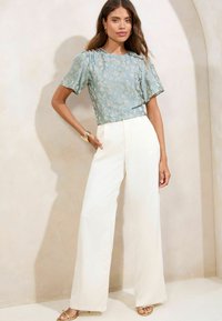Blusa a fiori in tonalità azzurro chiaro con maniche corte, abbinata a pantaloni larghi bianchi e sandali dorati. Tessuto morbido, vestibilità rilassata.