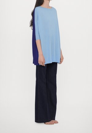Femme portant une tunique ample bleu clair avec un panneau arrière bleu foncé, un jean large foncé et des sandales ouvertes, debout de profil.