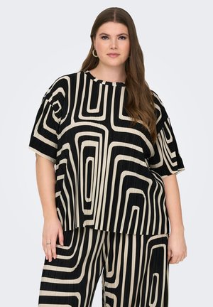 Kvinde med langt brunt hår iført sort og beige løstsiddende top med geometrisk mønster samt matchende bukser med vide ben mod en ensfarvet lys baggrund.