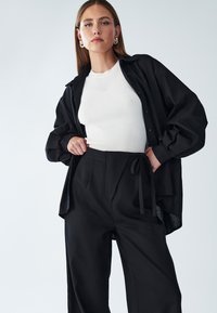 Zwarte oversized shirt, witte ribbeltop en zwarte high-waisted broek met een strik. Soepele materialen, relaxte pasvorm en eenvoudige styling.