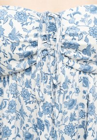 Λευκό ύφασμα με μπλε ανθισμένο και πεταλούδες, με σούρες στο κέντρο δεμένες με δύο λεπτές κορδέλες από το ίδιο ύφασμα.