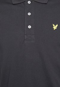 Czarny polo w kolorze ciemnoszarym, wykonany z bawełny, z plisą na trzy guziki oraz żółtym haftowanym logo ptaka po lewej stronie klatki piersiowej. Gładka faktura.