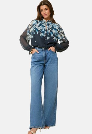 Black floral blouse with sheer sleeves and a tie neckline, paired with high-waisted wide-leg blue jeans. Sandals complete the look.
