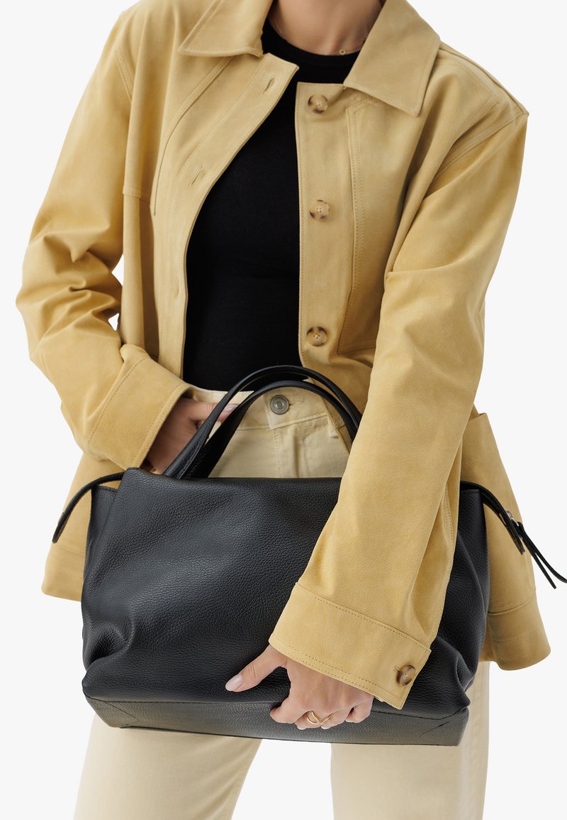 Sac à main en cuir noir avec surface texturée et poignées doubles, tenu contre une veste en daim jaune. Design simple sans motifs visibles.