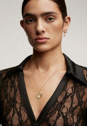 Mujer con blusa de encaje negro y cuello de satén, collar de pendiente dorado y pequeños pendientes en forma de aro en dorado y rojo, expresión neutral.