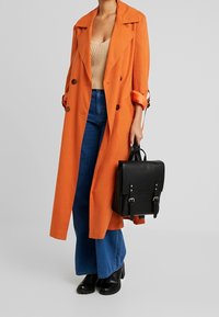 Cappotto arancione con ampi risvolti, chiusure a bottoni; maglia lavorata beige; jeans svasati blu; zaino in pelle nera con tracolle; stivaletti neri.