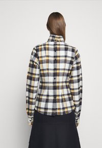 Checkered flannel shirt in white, navy, and mustard with a fitted cut. Features a pointed collar and button closures down the back.
