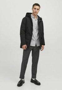 Zwarte geïsoleerde jas met een capuchon, ritssluiting en voorzakken, gedragen over een lichtgrijs button-up shirt en grijze broek.
