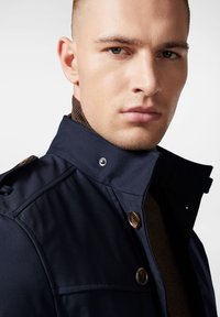 Granatowy płaszcz z wysokim kołnierzem i zapięciem na napy, z teksturowaną brązową podszewką i shoulder epaulettes.