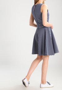 Femme portant une robe sans manches rayée bleue et blanche avec une jupe évasée et des baskets blanches avec des lacets noirs, se tenant sur une jambe.