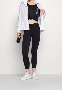 Svart atletisk crop top med knuten framtill, svarta högmidjade leggings, vit zip-up jacka med svarta ränder, och vita atletiska skor.