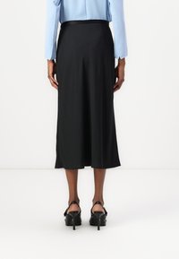 Svart midi skjørt med en jevn tekstur og subtil drapering, med en tettsittende midje og splitt på sidene, kombinert med svarte høyhælte sko.