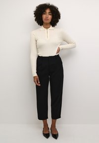 Top a maniche lunghe a coste color crema con colletto e zip a metà, abbinato a pantaloni neri a vita alta e scarpe basse a punta nera.