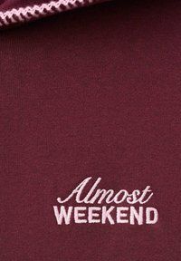 Tela marrón con texto bordado en rosa "Casi FIN DE SEMANA" y costura en zigzag rosa a juego a lo largo del borde.