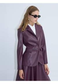 Blazer in finta pelle viola con silhouette aderente, rever a cancellino, due bottoni e vita con cintura; indossato sopra una gonna coordinata.