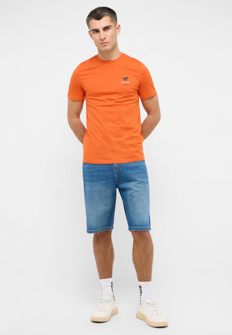 Pomarańczowa bawełniana koszulka z krótkim rękawem i logo na piersi, w połączeniu z niebieskimi szortami dżinsowymi i białymi trampkami.