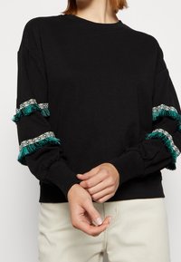 Czarna bluza z trzema czwartymi rękawami z tealowymi frędzlami i tkanym detalem przy mankietach. Miękka tkanina, luźny krój.