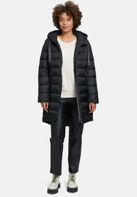 Zwarte donsjas met capuchon, horizontaal gestikt ontwerp, sluiting met ritssluiting aan de voorkant, twee zijzakken, gedragen over een lichte trui en zwarte broek.