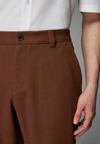 Ruske priređene hlače od teksturiranog materijala, s crnim dugmetom, bočnim džepovima i petljom za pojas, prikazane s belom košuljom.