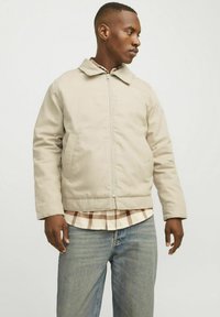 Beige, isoleret jakke med lynlås, rund krave og to sidelommer, båret over en ternet skjorte og parret med lyseblå jeans.
