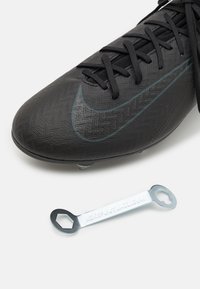 Botte de football noire avec une tige texturée, dotée de lacets et d'un swoosh contrasté. Accompagnée d'un outil en métal avec une extrémité hexagonale pour l'entretien.
