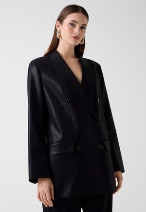 Jeune femme aux cheveux bruns raides portant de grandes boucles d'oreilles en anneau et un blazer noir à double boutonnage, sur un fond clair uni.