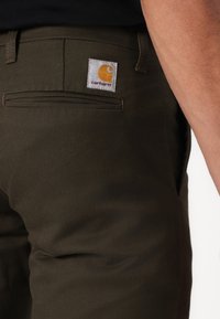 Olivengrønne bukser lavet af slidstærkt stof, med en baglomme der har et kvadratisk Carhartt mærke i hvidt og orange. Skræddersyet pasform med synlige stikninger.