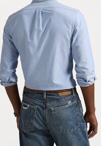 Ljusblå skjorta med knappar och uppvikta ärmar, med en veckad detalj baktill. Bärs med slitna blå jeans och ett brunt bälte.