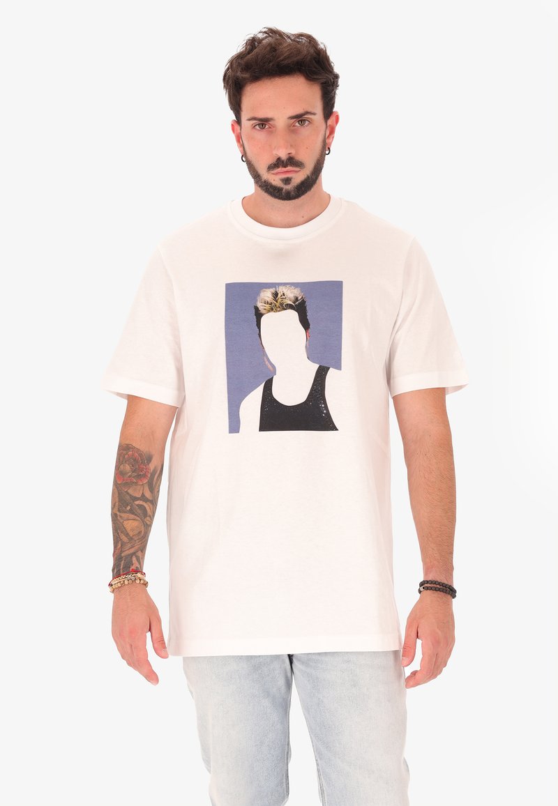 Homme avec une barbe et des tatouages portant un T-shirt blanc avec un imprimé graphique sans visage et un jean bleu clair, se tenant devant un fond uni.