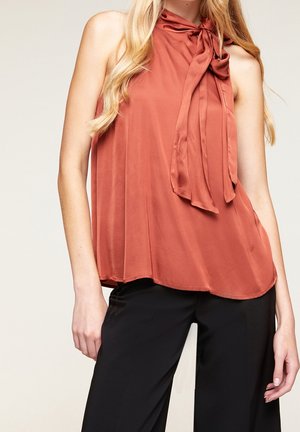 Vrouw met een mouwloze, roestkleurige satijnen blouse met een gestrikte strik bij de hals en zwarte broek, weergegeven van schouders tot heupen.