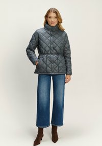 Žena oblečená v šedej prešívanej bunde s nastaviteľným pásom, modrých širokých džínsoch a hnedých čižmách na podpätku, stojí pred jednoduchým pozadím.