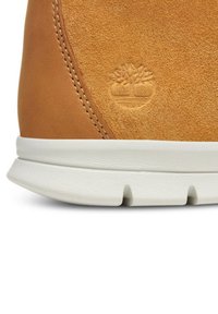 Sneaker aus beige Wildleder und Leder mit einer weißen Gummisohle, genähten Akzenten und prägedem Baumlogo. Weiche Textur und runde Form.