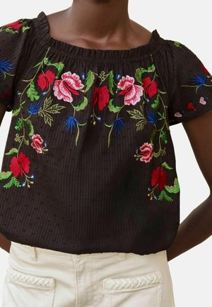 Blouse noire texturée avec des fleurs et des feuilles brodées multicolores, associée à un pantalon clair avec des poches tressées.