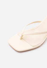 Beżowy sandał z kwadratowym czubkiem, cienkimi paskami i minimalistycznym wzorem na białym tle.