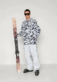 Egy snowboardos mintás sötétkék és fehér kapucnis pulóverben, világosszürke bő nadrágban és fekete sícsizmában áll, miközben egy színes snowboardot tart.