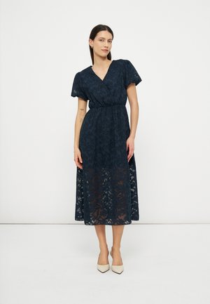 Γυναίκα φορώντας midi φόρεμα από μπλε δαντέλα με φουσκωτά μανίκια και λαιμόκοψη σε σχήμα V, συνδυασμένο με μπεζ μυτερές γόβες, στέκεται μπροστά σε απλό λευκό φόντο.